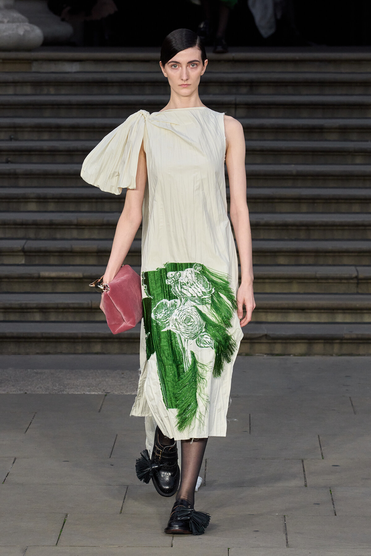 Model on stairs wearing a cream, one-shoulder dress with a bold green floral print at the hem, carrying a pink clutch and wearing black tassel loafers.