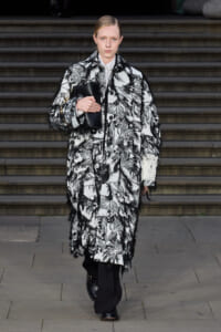 Model walking down stairs in a black-and-white botanical coat with fringe, clutch held under her arm.