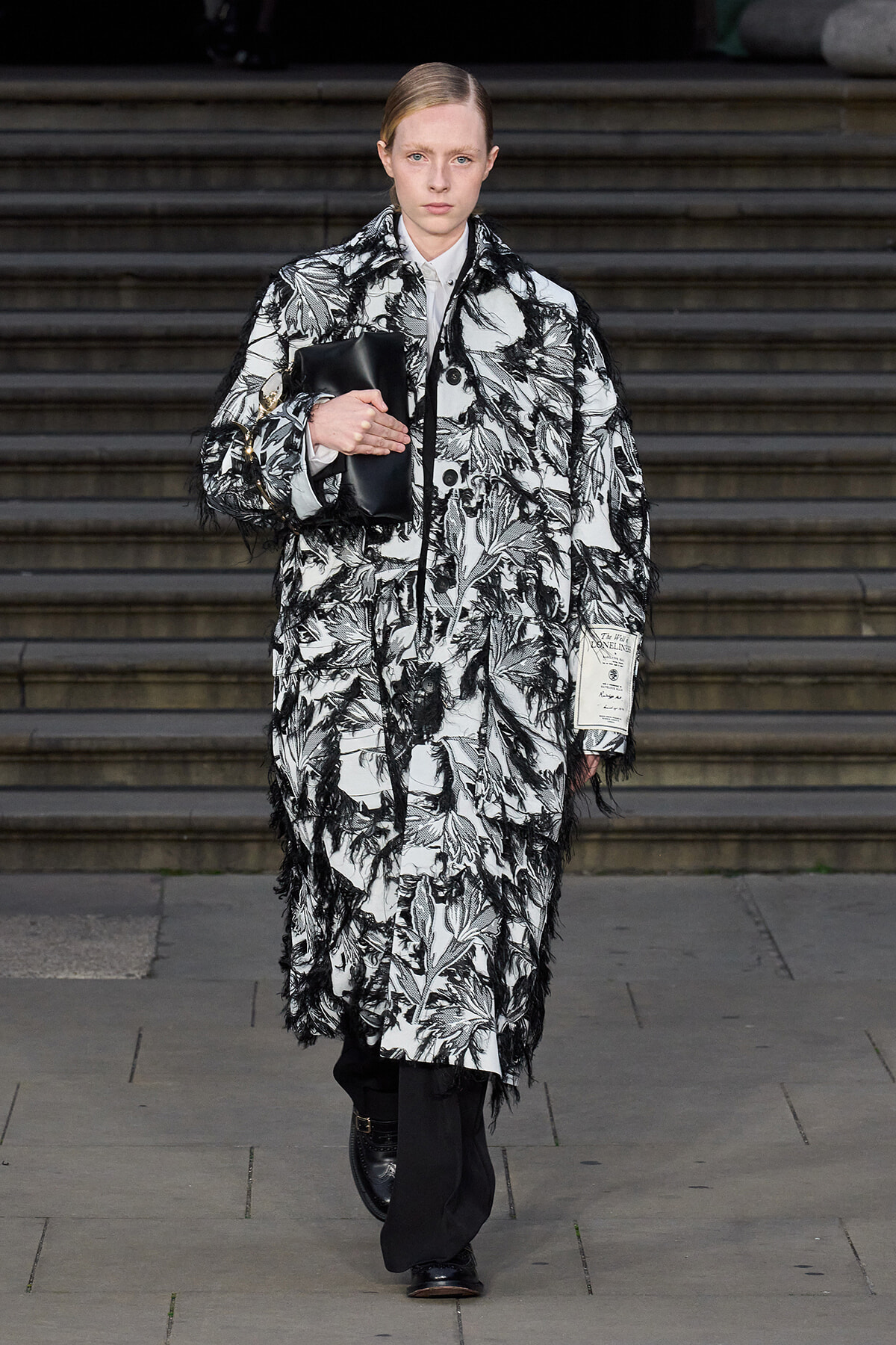 Model walking down stairs in a black-and-white botanical coat with fringe, clutch held under her arm.