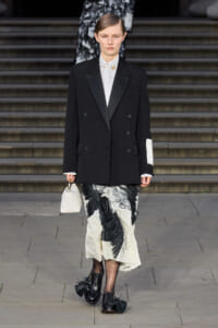 Model walking down stairs in a black double-breasted blazer, white shirt, and a black-and-white floral midi skirt, carrying a small white handbag.