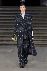 Model walking in a navy pinstripe double-breasted suit with small floral embroidery, paired with matching wide-leg pants and a lavender lining, carrying an olive green clutch.