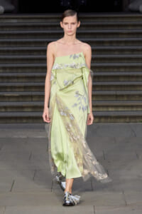 Model on a runway wearing a pale lime-green slip dress with silver floral embroidery and a sheer overlay.