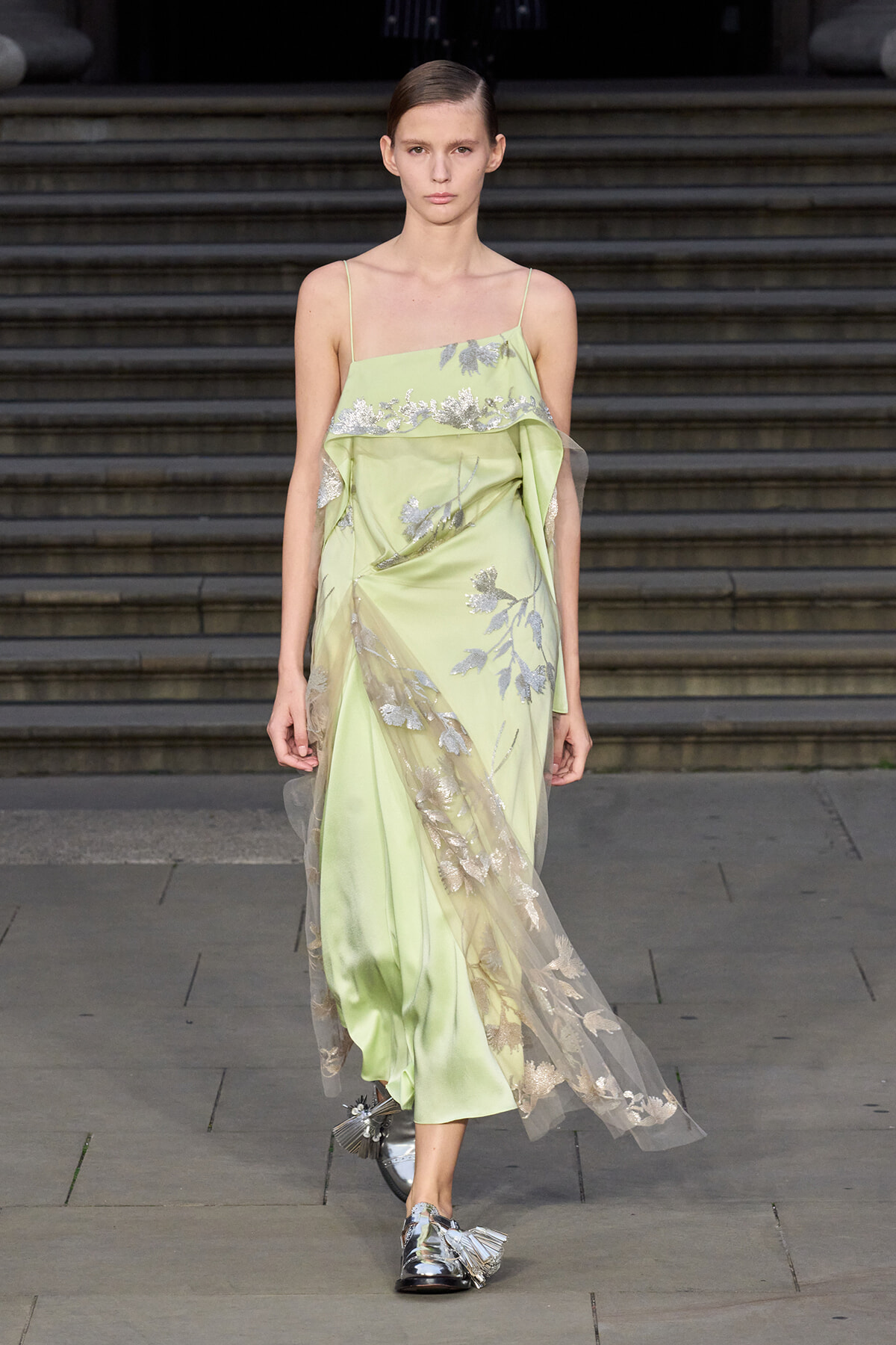 Model on a runway wearing a pale lime-green slip dress with silver floral embroidery and a sheer overlay.