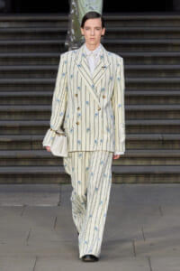 Model wearing a cream striped double-breasted suit with blue floral embroidery, white shirt, matching wide-leg pants, and a small white handbag while walking down steps.