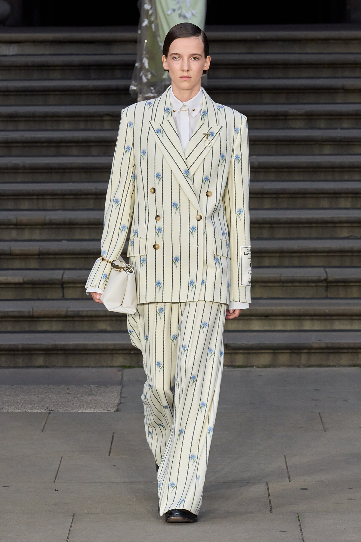 Model wearing a cream striped double-breasted suit with blue floral embroidery, white shirt, matching wide-leg pants, and a small white handbag while walking down steps.