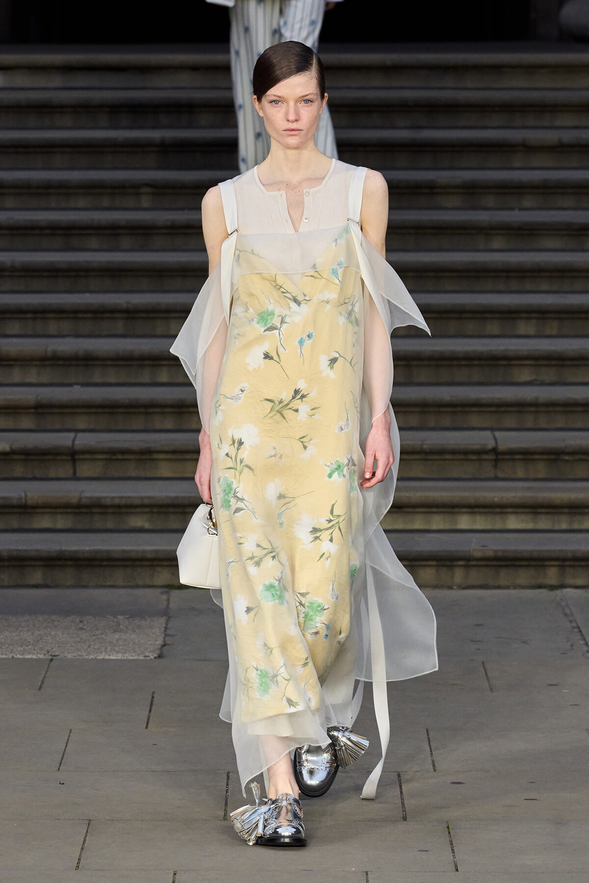 Model on a runway wearing a pale yellow floral slip dress with a sheer cape and light-colored top, paired with silver tassel loafers and a small white handbag on steps behind.