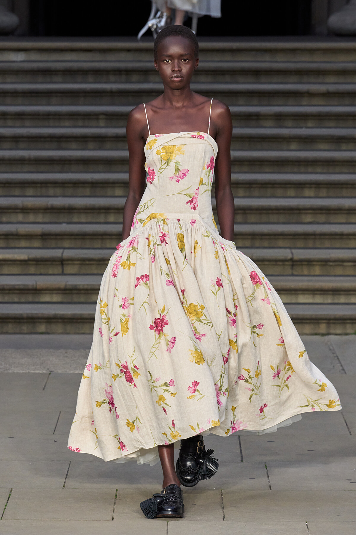 Model walking a runway in a cream floral-print maxi dress with pink and yellow flowers and thin straps, paired with black tassel shoes