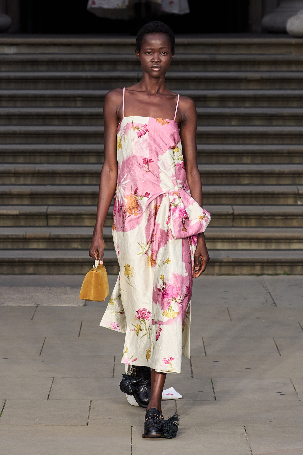 Model walking a runway in a pink and cream floral midi dress with thin straps, holding a small mustard handbag.