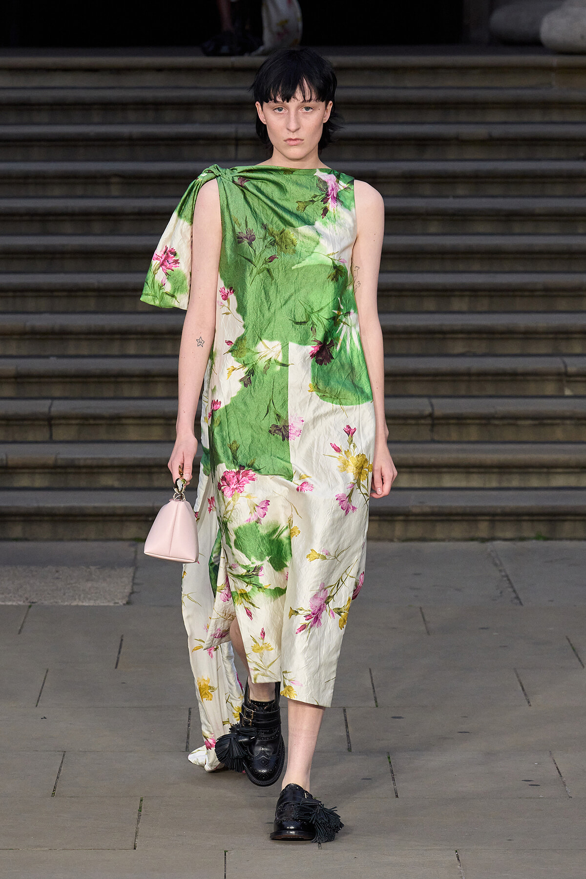 Model walking down stone steps in a green and cream floral midi dress with a draped cape over one shoulder, carrying a small pink handbag and wearing black fringe loafers.