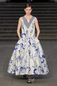 Runway model in a white and blue floral ball gown with a large waist bow, worn over a grey sleeveless top; silver shoes visible.
