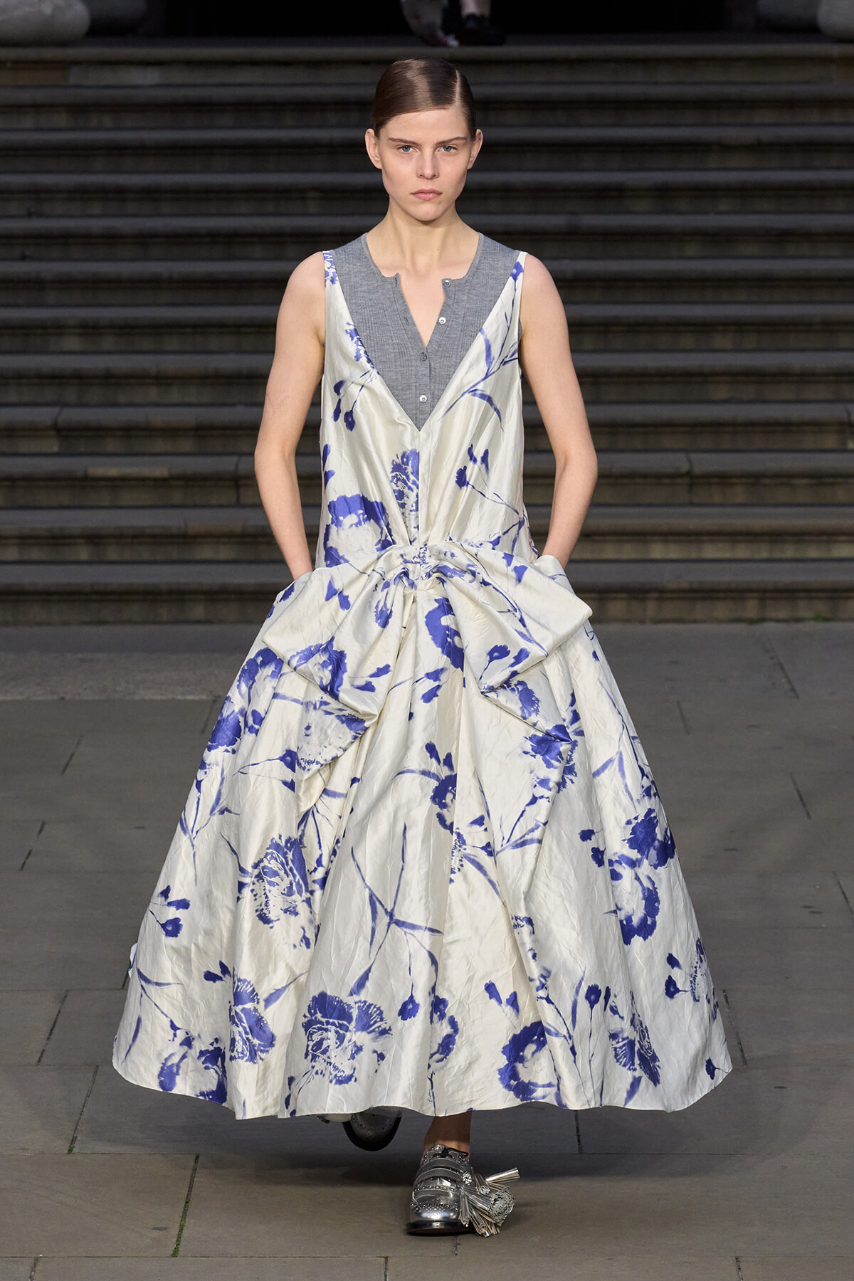 Runway model in a white and blue floral ball gown with a large waist bow, worn over a grey sleeveless top; silver shoes visible.