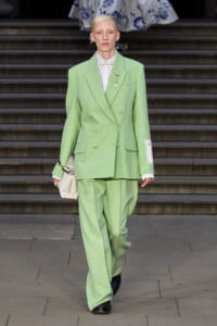 Model wearing a mint green double-breasted suit with wide-shouldered blazer and matching wide-leg trousers, white shirt, gold lapel pin, and carrying a small white bag while walking down steps.