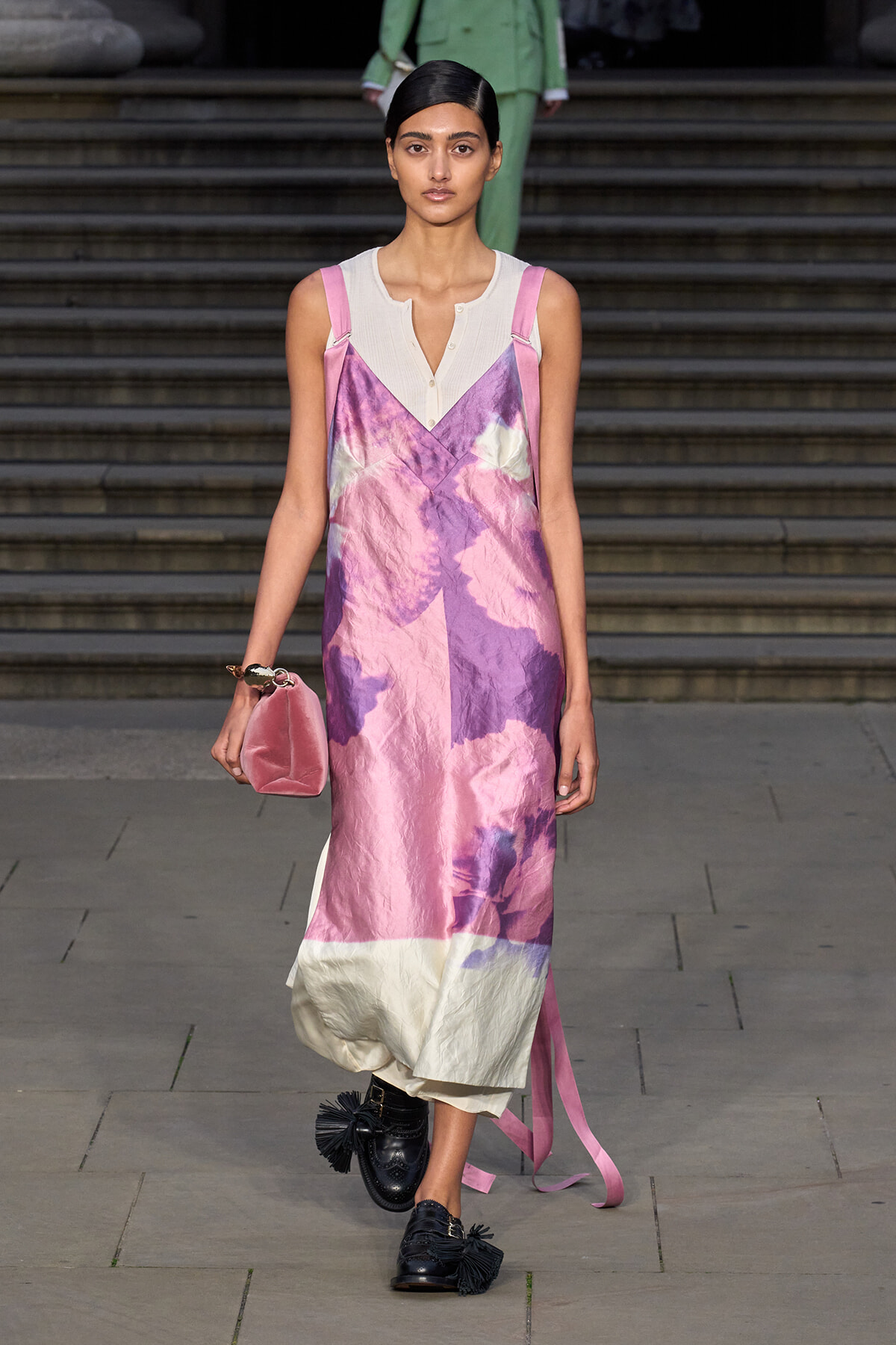 Model walking runway in pink–purple tie-dye slip dress over white top, holding a small pink clutch; black tasseled loafers visible.