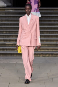 Model on a runway wearing a pastel pink double-breasted suit with a white shirt, carrying a bright yellow clutch down the stairs.