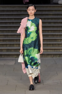 Runway model in a sleeveless green and blue abstract print dress with a pink wrap over one shoulder and a small white handbag.