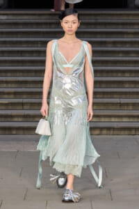 Model walking down steps in a pale mint metallic gown with sheer panels and matching ribbons, carrying a small white handbag, silver shoes visible at the bottom.