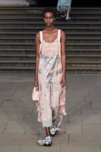 Female model walks a runway in a pale pink lace dress with metallic panels and pink straps, carrying a small pink handbag.