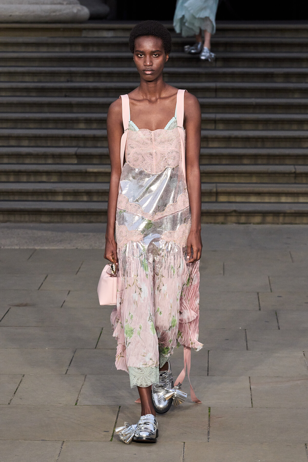 Female model walks a runway in a pale pink lace dress with metallic panels and pink straps, carrying a small pink handbag.