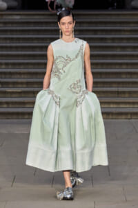 Model walking on a runway wearing a pale mint-green sleeveless gown with silver beaded embroidery and a voluminous skirt, paired with metallic loafers.
