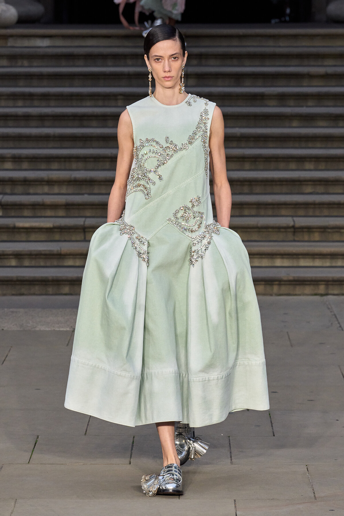 Model walking on a runway wearing a pale mint-green sleeveless gown with silver beaded embroidery and a voluminous skirt, paired with metallic loafers.