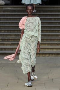 Model walks a runway in a pale mint lace dress with a pink draped sash and silver tassel shoes.