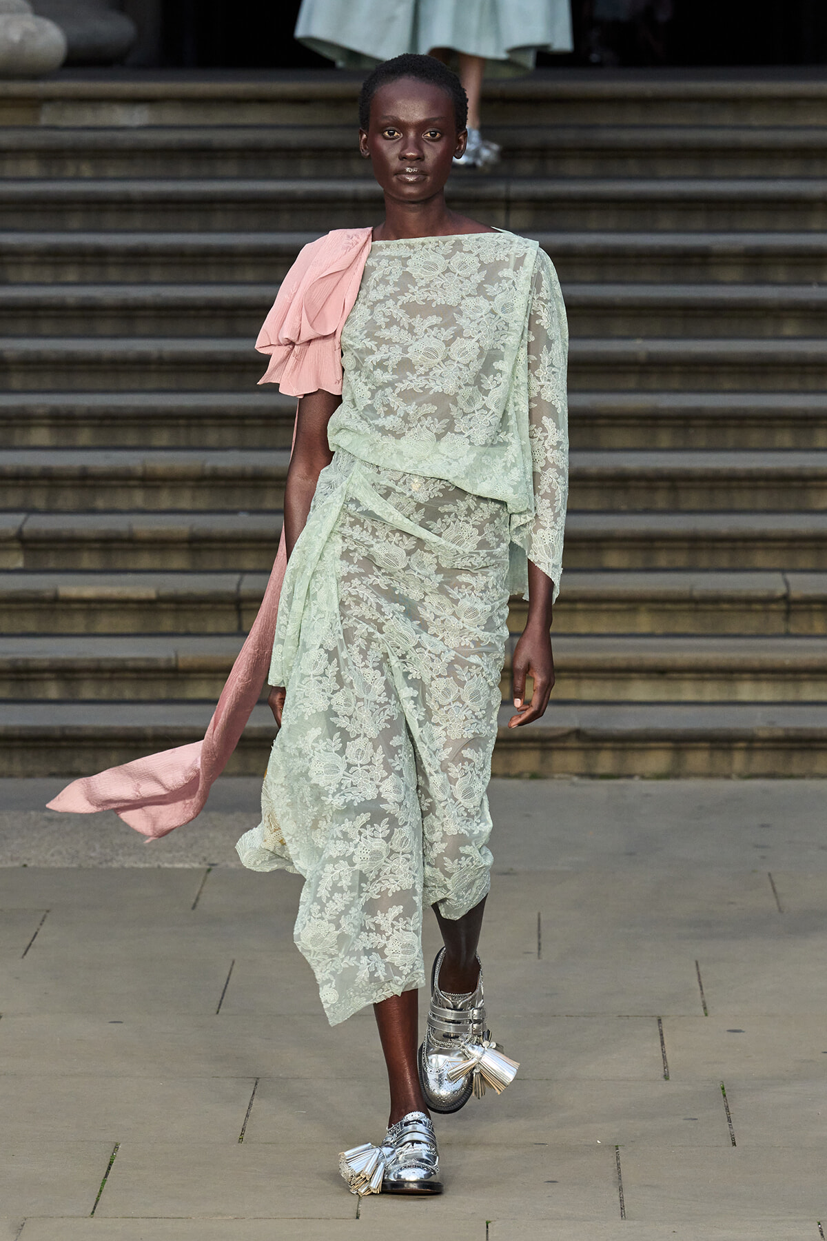 Model walks a runway in a pale mint lace dress with a pink draped sash and silver tassel shoes.