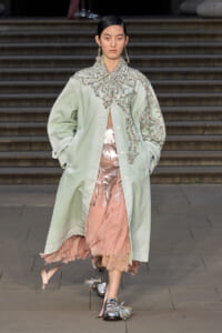 Runway model in a pale green oversized coat with silver bead embroidery over a pink sheer dress and metallic shoes.