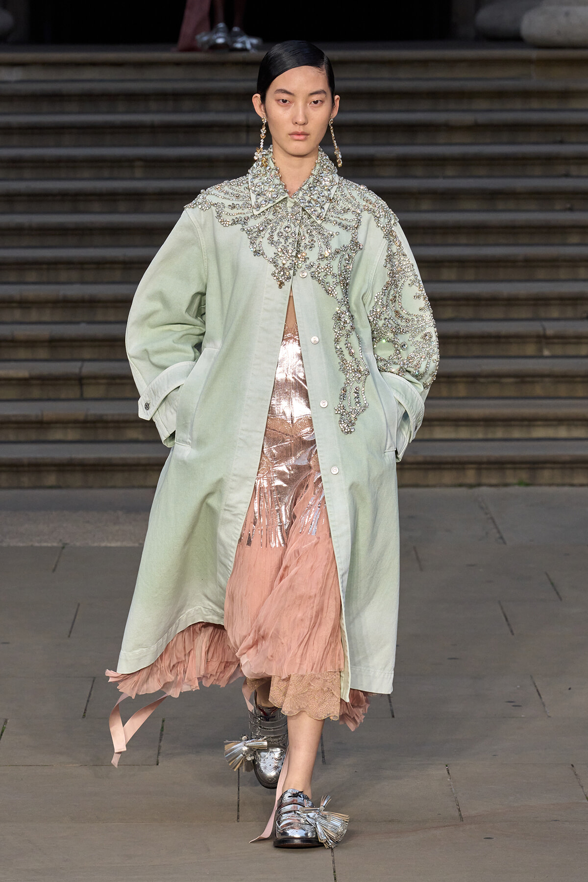 Runway model in a pale green oversized coat with silver bead embroidery over a pink sheer dress and metallic shoes.