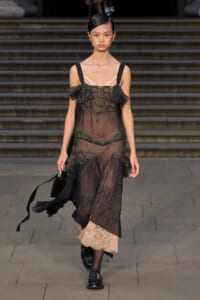 Runway model in a sheer black lace dress with gold embroidery, pink neckline underlay, walking with a black handbag on a stone steps backdrop.