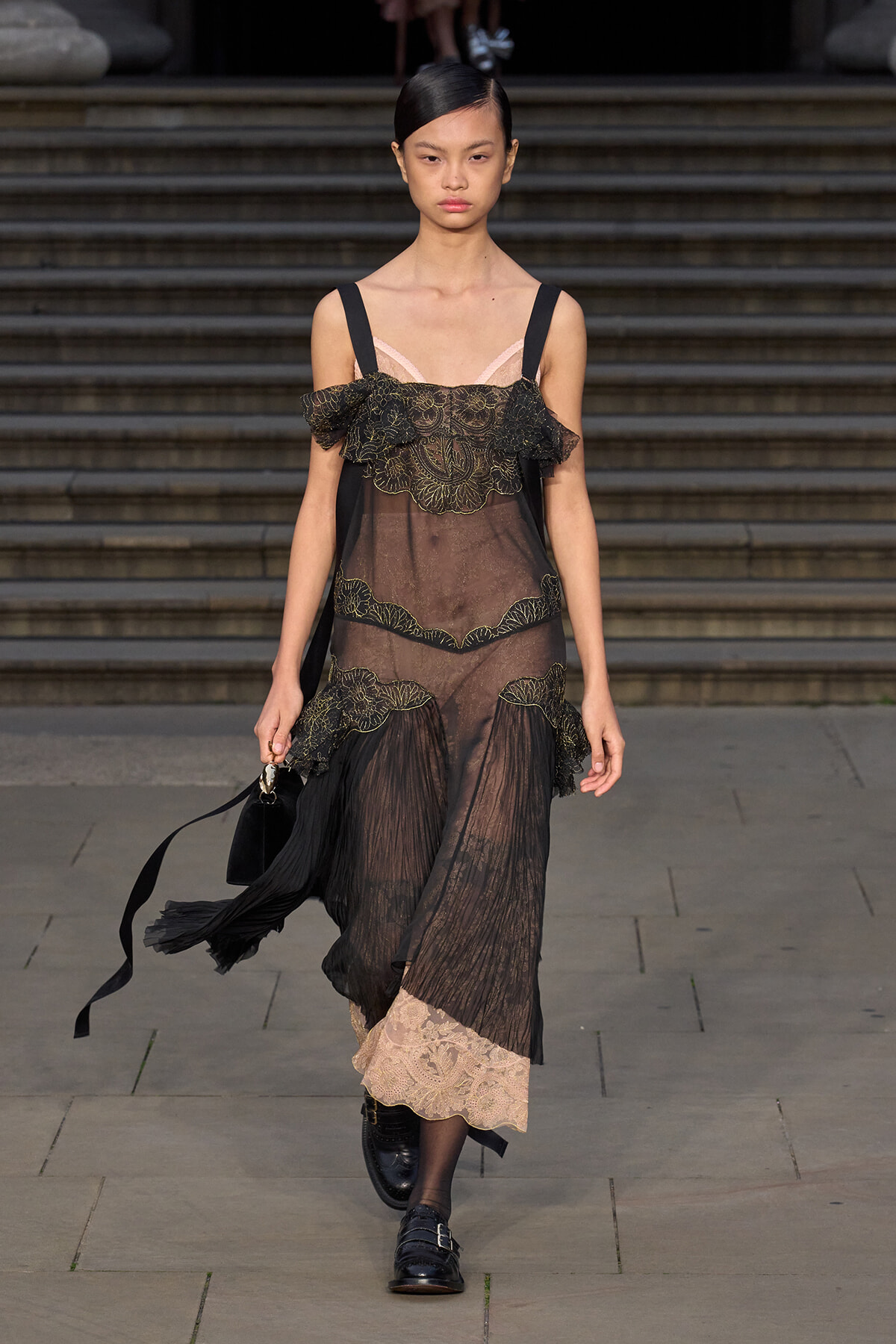 Runway model in a sheer black lace dress with gold embroidery, pink neckline underlay, walking with a black handbag on a stone steps backdrop.