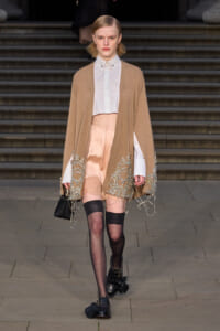 Model on runway wearing a tan cape with silver bead embroidery over a white cropped shirt and peach satin mini skirt, sheer black tights, tassel loafers, and a small black handbag.
