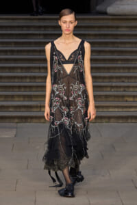 Model walking a black sheer embroidered dress with pink floral details on a runway.