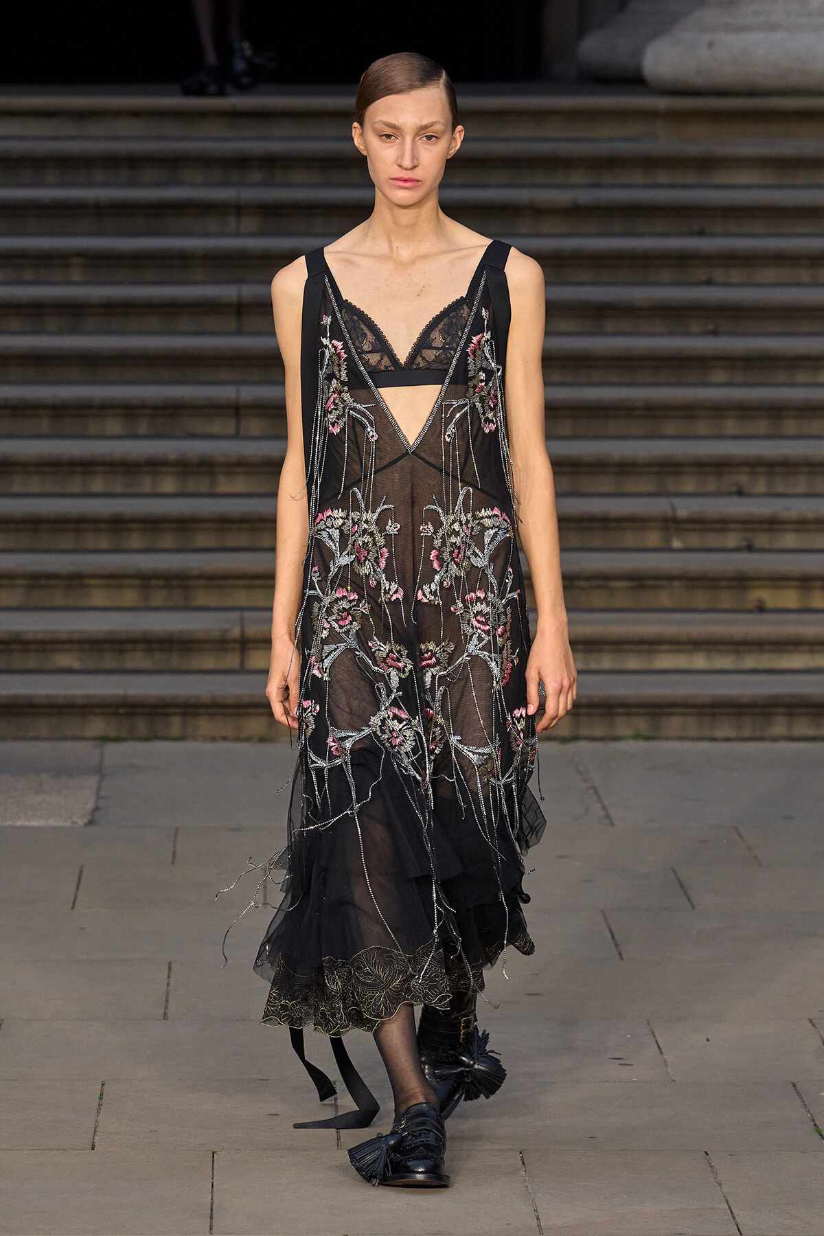 Model walking a black sheer embroidered dress with pink floral details on a runway.