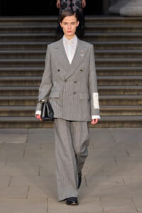 Model walking in a gray pinstripe double-breasted suit with white shirt, holding a black clutch on a runway.