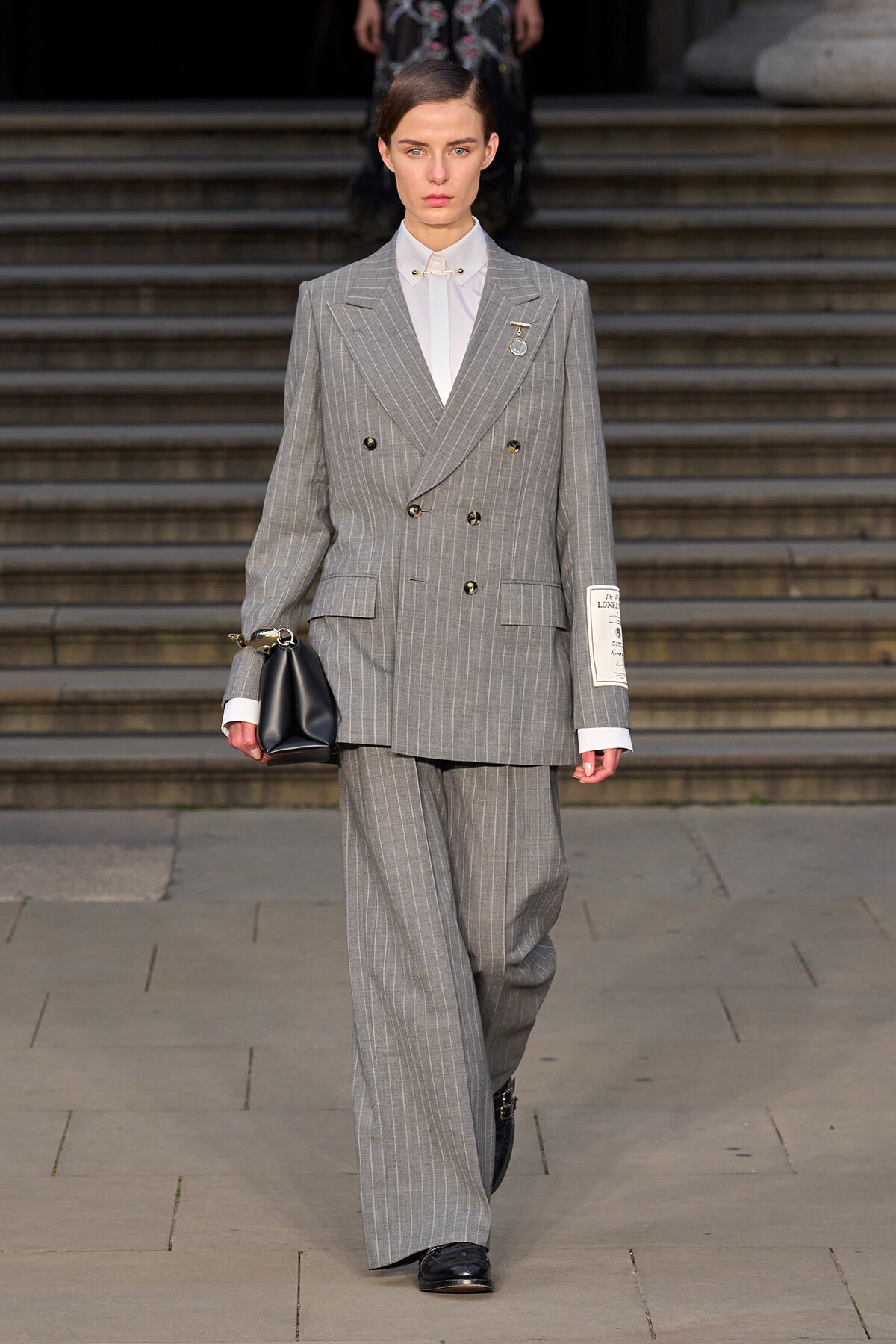 Model walking in a gray pinstripe double-breasted suit with white shirt, holding a black clutch on a runway.