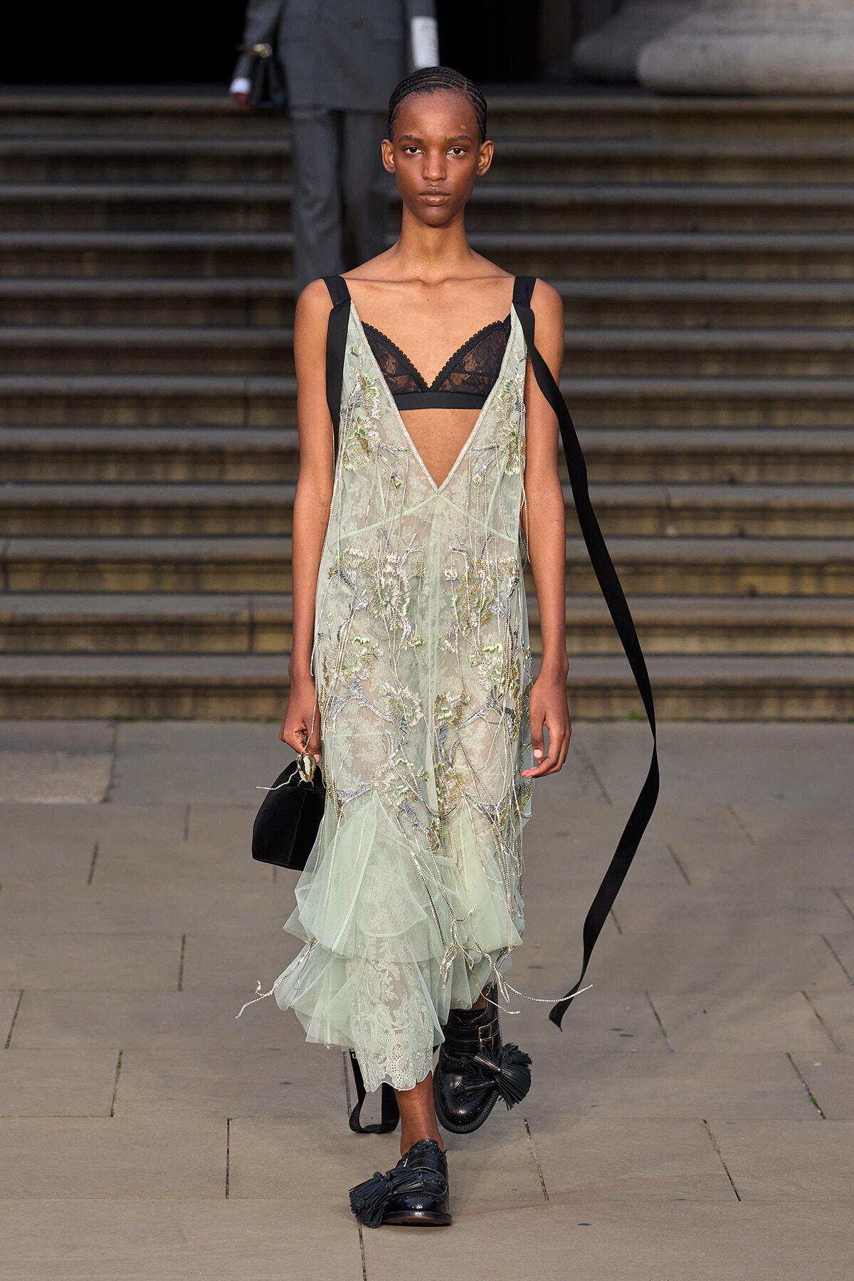 Model walking a runway in a pale mint sheer gown with silver embroidery over a black lace bra, holding a small black handbag.