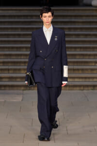 Female model in a navy pinstripe double-breasted suit with a white shirt and badge, carrying a black clutch, walking down steps.