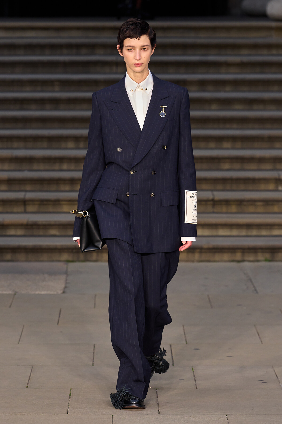 Female model in a navy pinstripe double-breasted suit with a white shirt and badge, carrying a black clutch, walking down steps.