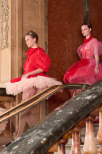 Two models in red and pink avant-garde gowns pose on a grand marble staircase, one in a voluminous red dress with layers of tulle and the other in a pink sheer gown with red beaded collar.