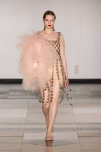 Runway model in a blush pink gown with a large tulle shoulder and gold floral chain appliqués along the bodice and skirt.