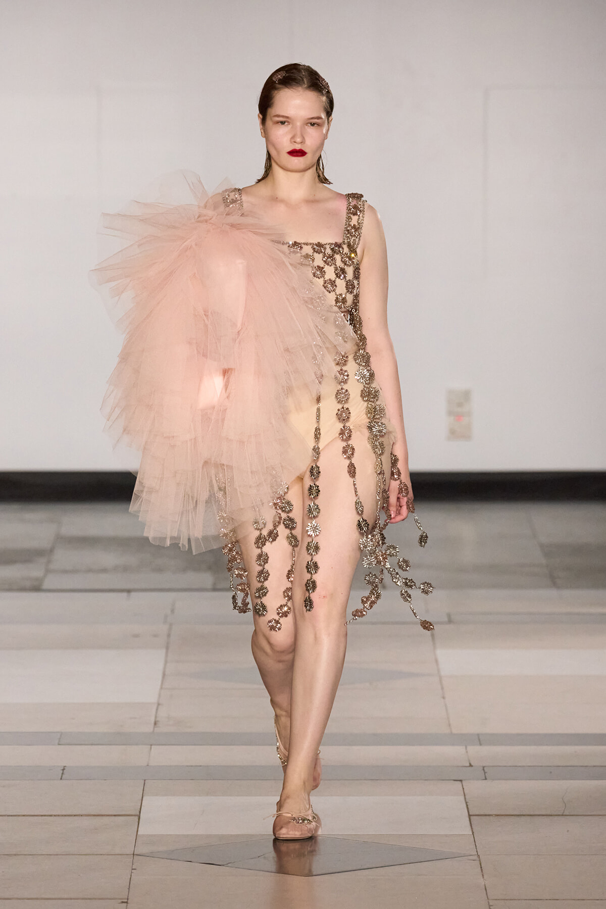 Runway model in a blush pink gown with a large tulle shoulder and gold floral chain appliqués along the bodice and skirt.