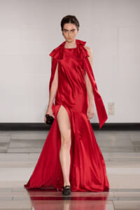 Model wearing a red satin gown with a high slit and cape-like draped sleeves on a runway, holding a small black clutch