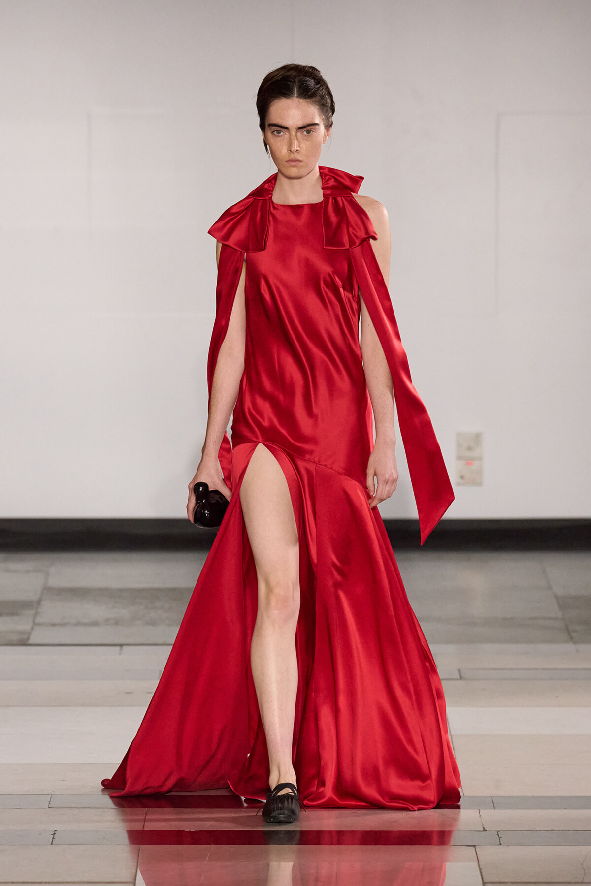 Model wearing a red satin gown with a high slit and cape-like draped sleeves on a runway, holding a small black clutch