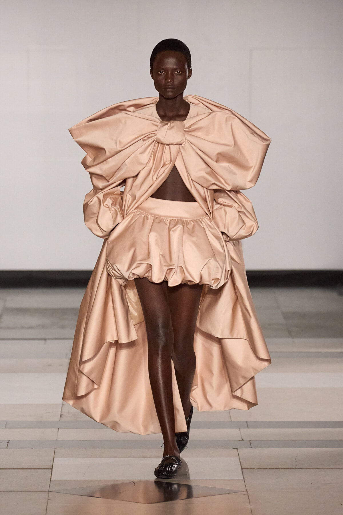 Model walks the runway in a peach satin ensemble with a voluminous bowed top and bubble skirt.