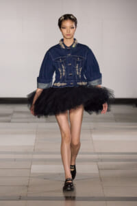 Model walking a runway in a dark denim jacket with silver embroidery and a voluminous black tulle skirt, black loafers.