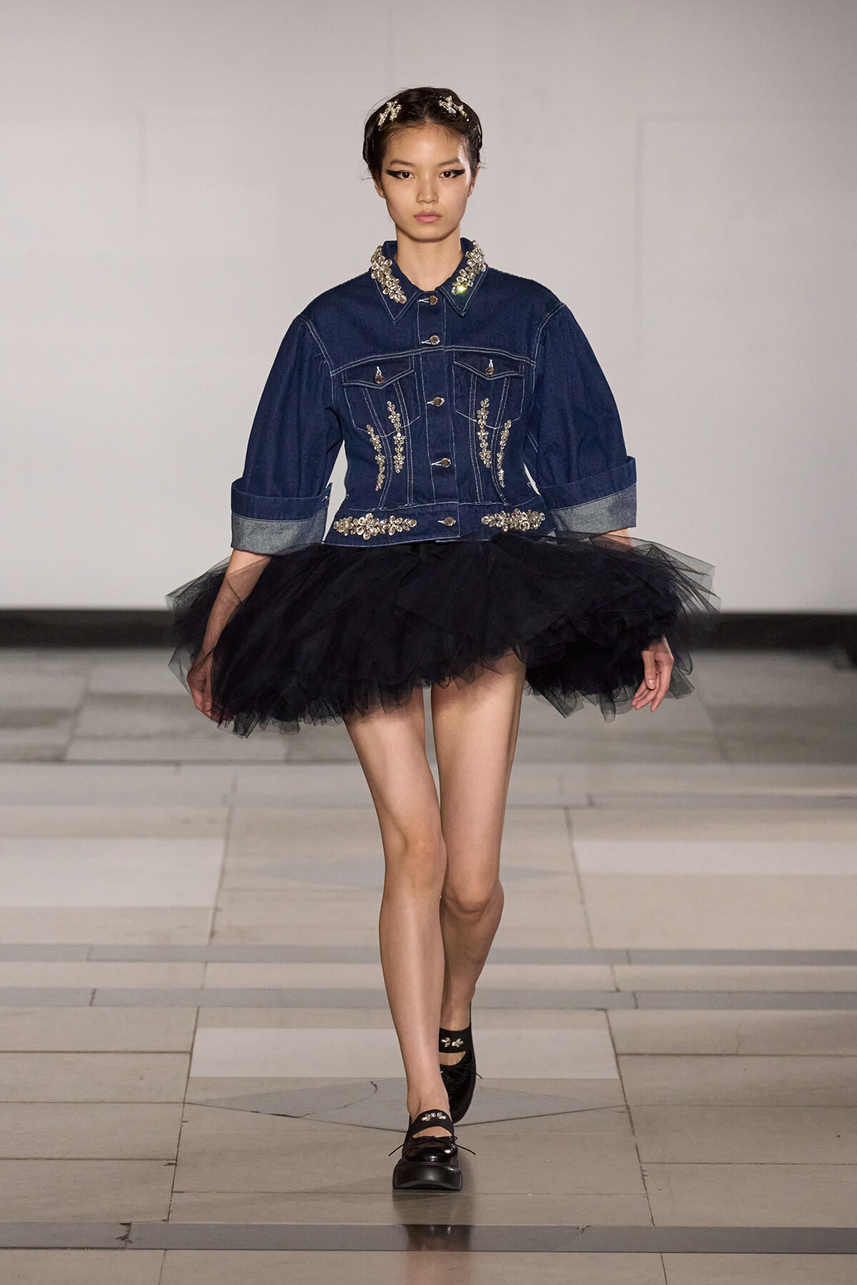 Model walking a runway in a dark denim jacket with silver embroidery and a voluminous black tulle skirt, black loafers.