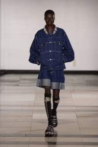 Male model on a runway wearing an oversized dark denim jacket with pearl-embellished collar and matching denim shorts, plus ornate knee-high socks and jeweled sandals.