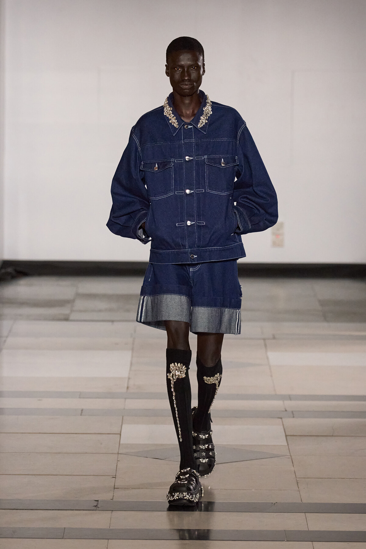 Male model on a runway wearing an oversized dark denim jacket with pearl-embellished collar and matching denim shorts, plus ornate knee-high socks and jeweled sandals.