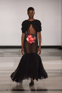 Model on a runway wearing a black sheer gown with a cropped top, exposed midriff, red floral panel, and a voluminous tulle mermaid skirt.