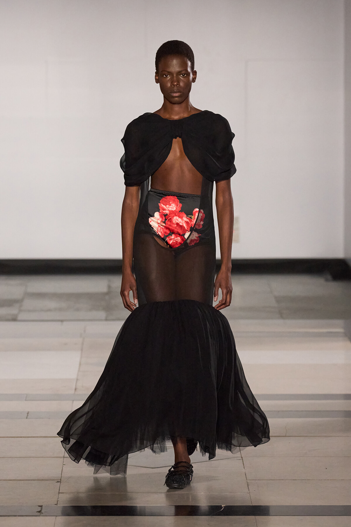 Model on a runway wearing a black sheer gown with a cropped top, exposed midriff, red floral panel, and a voluminous tulle mermaid skirt.